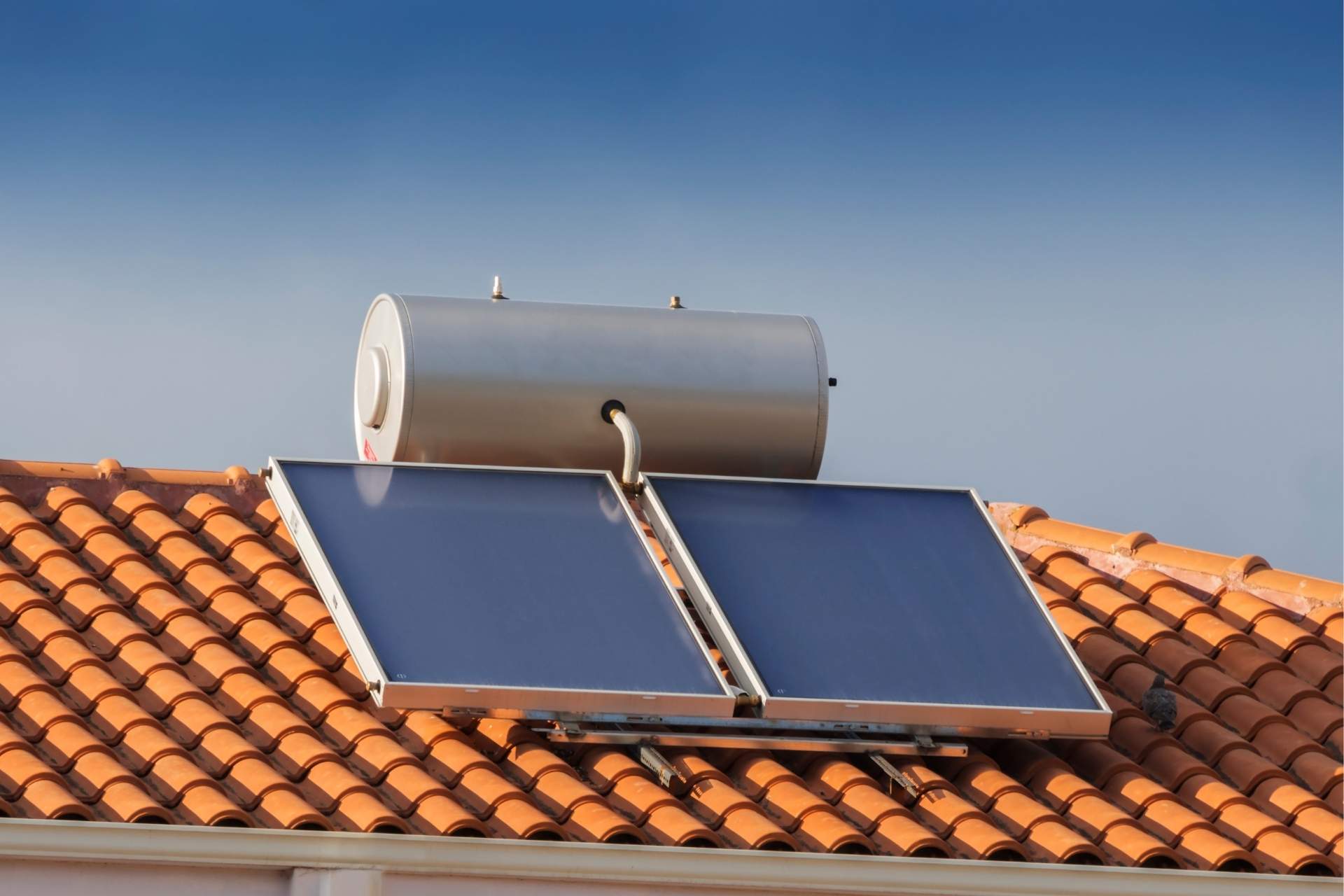 Image montrant des capteurs solaires thermiques Ulticom installés sur le toit d'un bâtiment. Les capteurs, de couleur bleu foncé, sont alignés sur le toit en tuiles orange. Un gros réservoir de stockage d'eau chaude solaire est également visible sur le toit. L'image illustre l'utilisation de l'énergie solaire thermique pour produire de l'eau chaude dans cette maison.