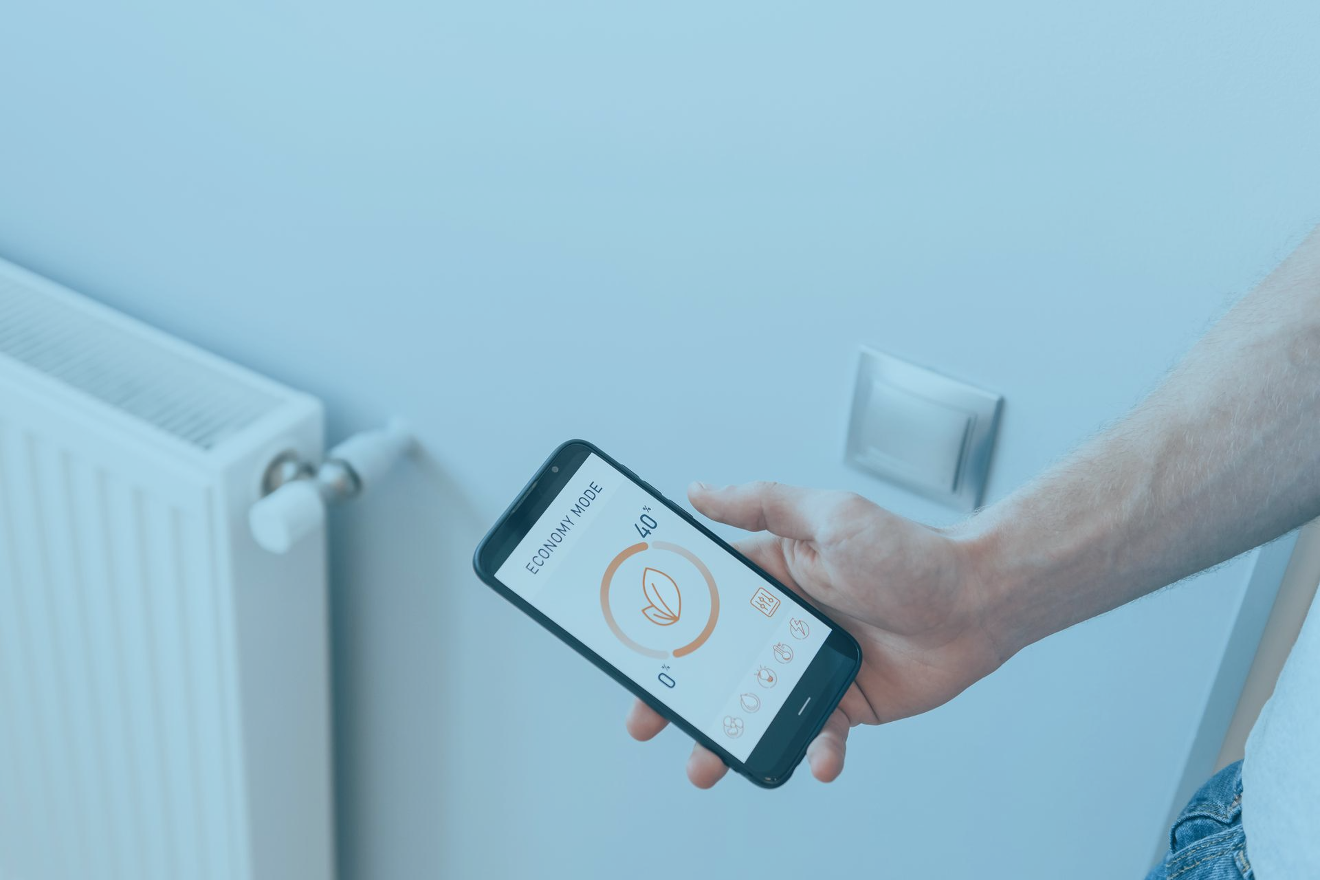 Une personne à côté d'un radiateur électrique sur lequel est installé un thermostat connecté, tenant en main un smartphone sur lequel est affiché une application pour contrôler la température du radiateur pour réaliser des économies sur ses factures d'énergies